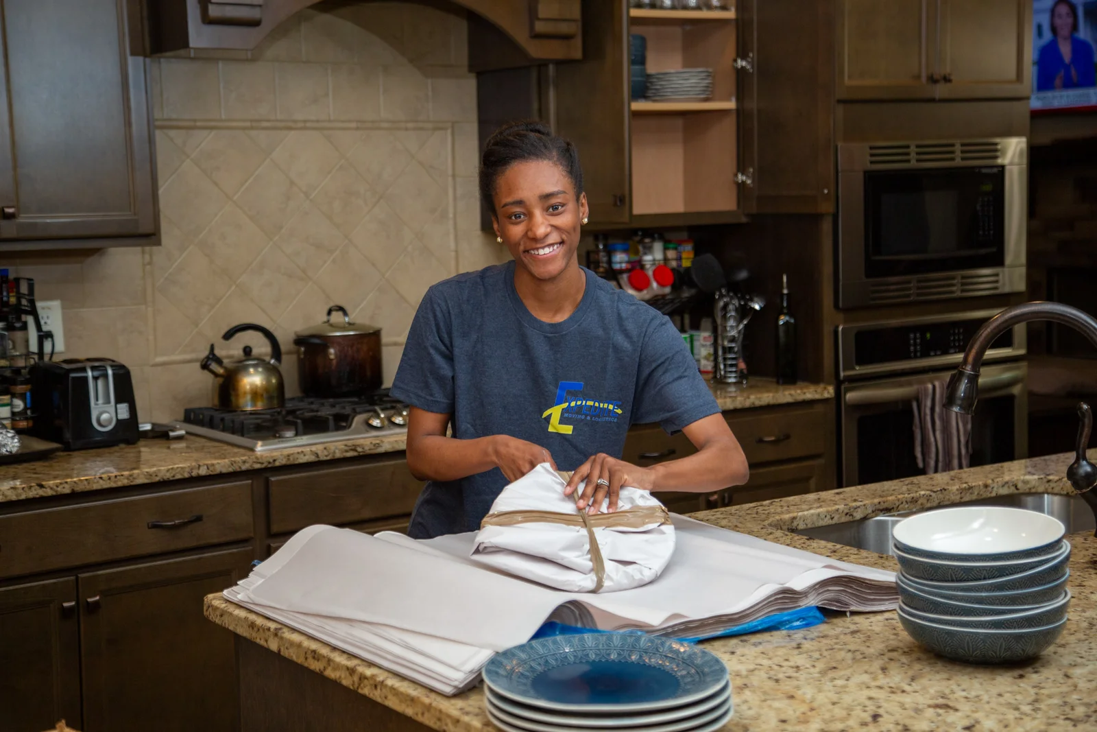 A professional mover smiling while carefully wrapping and packing dishes during a move.