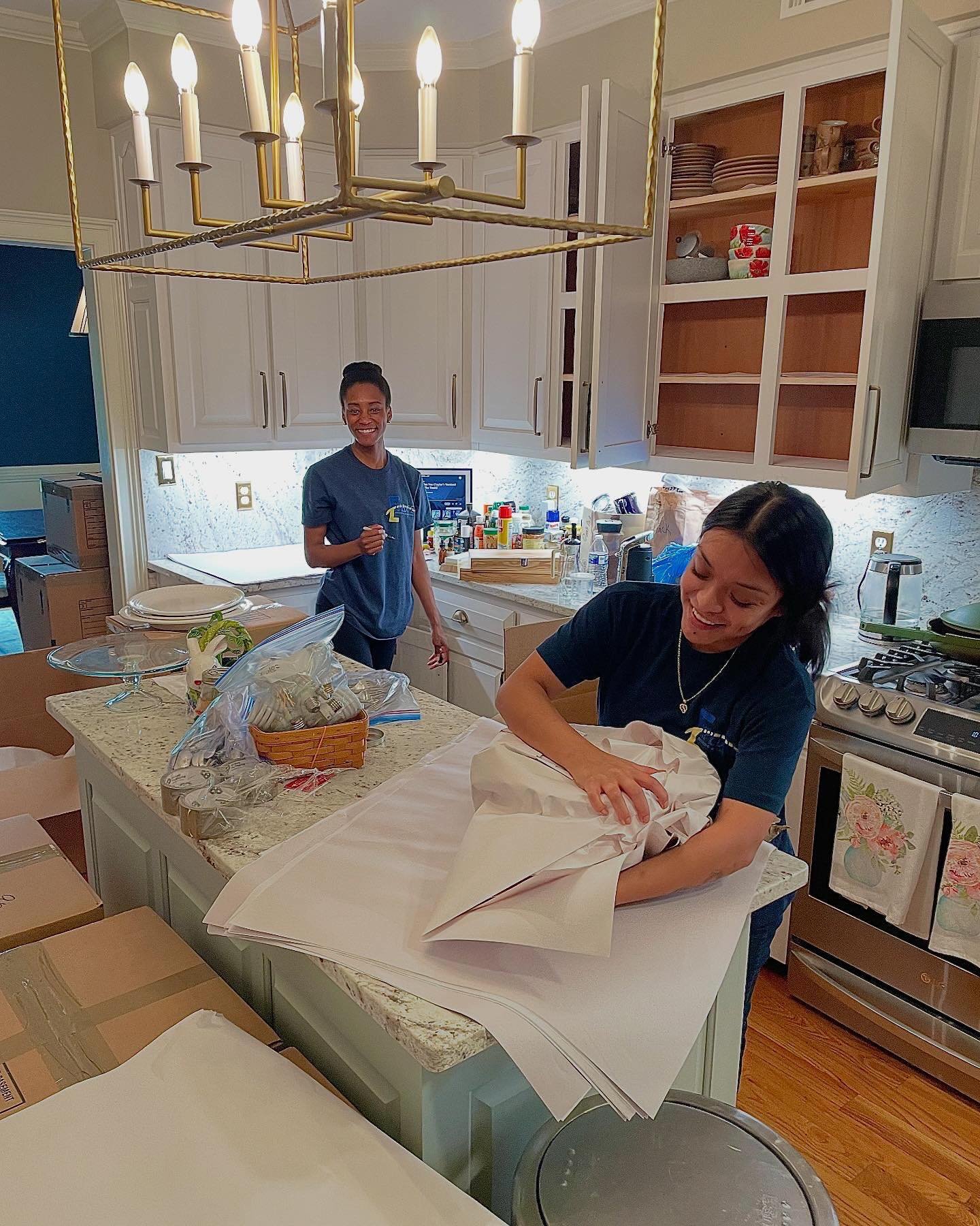 Two professional movers in a kitchen wrapping and packing stacked plates with packing paper for relocation.